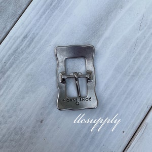May include: Silver metal belt buckle with a rectangular shape and a wavy edge. The buckle has the words "HORSE SHOE" engraved on it.