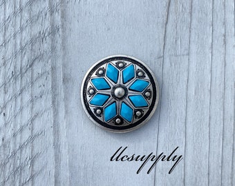 May include: A silver and turquoise round concho with a starburst design. The concho is on a light wood background.