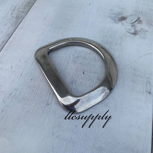 May include: A silver metal D-ring hardware, a metal loop with a D-shape, on a white wooden surface.