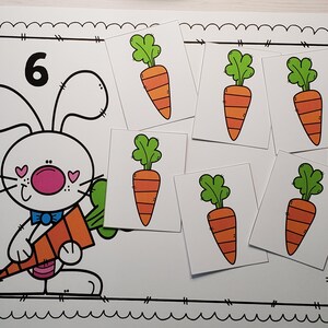 May include: A white bunny with pink cheeks and a blue bow tie holds a large orange carrot. Six smaller orange carrots are on white cards with green tops. The number 6 is written in black on a white card.