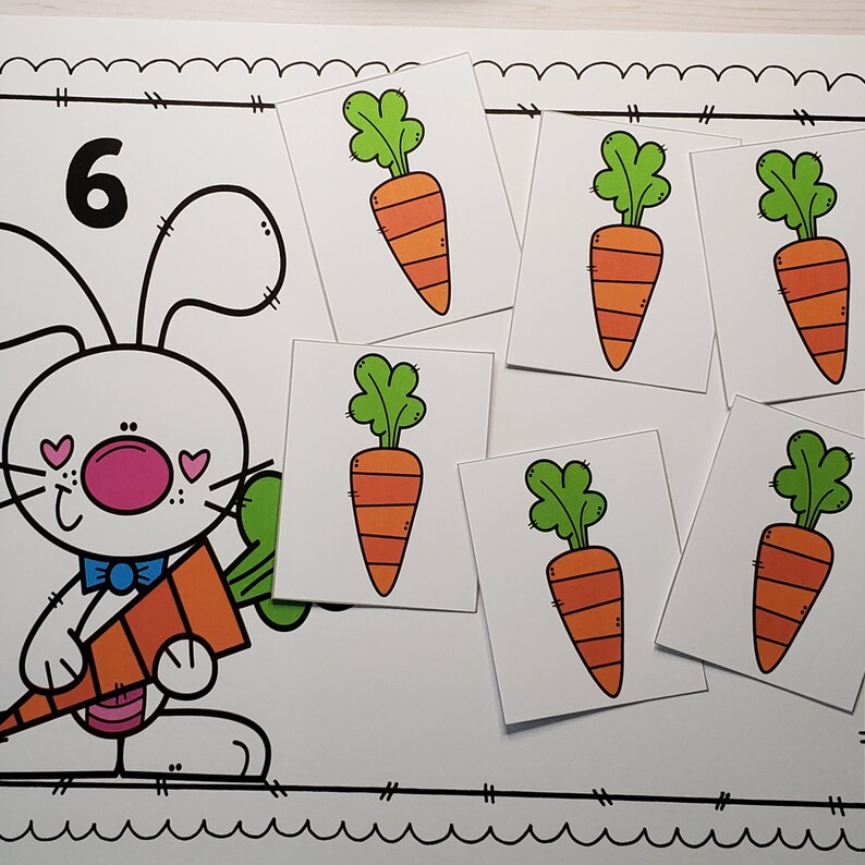 May include: A white bunny with pink cheeks and a blue bow tie holds a large orange carrot. Six smaller orange carrots are on white cards with green tops. The number 6 is written in black on a white card.