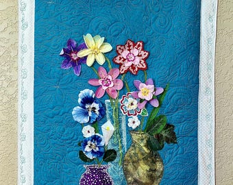 May include: A rectangular fabric wall hanging featuring a quilted teal background with a swirling pattern. The design displays three vases containing various colourful fabric flowers in shades of purple, yellow, red, and blue, framed by a white border.