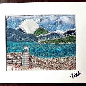 May include: A framed artwork depicting a lighthouse on a beach with a full moon and mountains in the background. The artwork is made with fabric and embroidery.