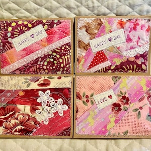 May include: Four rectangular greeting cards featuring fabric collage designs in shades of pink, red, and white. Two cards display the text HAPPY DAY with a heart icon, one shows the text LOVE with a heart, and one features white lace flowers.