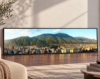 May include: A panoramic rectangular wall art print featuring a high-resolution landscape photograph of a dense city skyline nestled at the base of a large, green, sunlit mountain range under a clear blue sky with scattered white clouds.