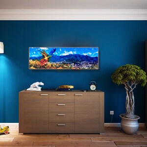 May include: A colorful abstract painting of a bird flying over a city landscape. The painting is framed and hanging on a blue wall in a room with a wooden dresser, a potted plant, and a bookshelf.