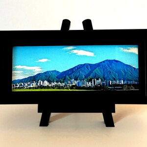 May include: A black framed picture with a cityscape and mountain range in the background. The picture is on a small easel.