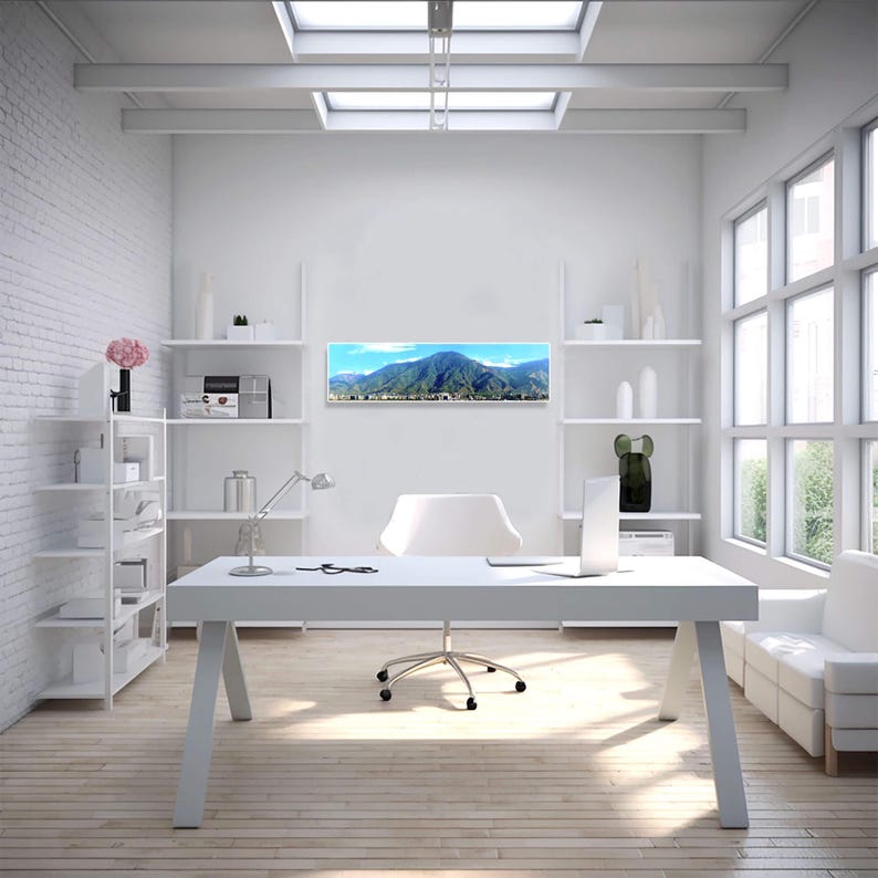 May include: A white office desk with a white chair and a white bookshelf. The desk has a white lamp and a laptop on it. The wall behind the desk has a panoramic photo of a mountain range.