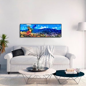 May include: A colorful abstract painting of a bird flying over a mountain range and a city in the distance. The painting is framed in black and hung on a white wall.