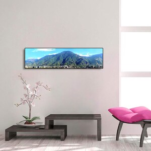 May include: A panoramic photograph of a mountain range with a city in the foreground. The photo is framed in a black frame and is hanging on a white wall.