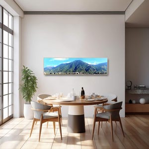 May include: A panoramic photograph of a mountain range with a city in the foreground. The photograph is framed in a gold frame and is hanging on a white wall above a dining table with chairs.