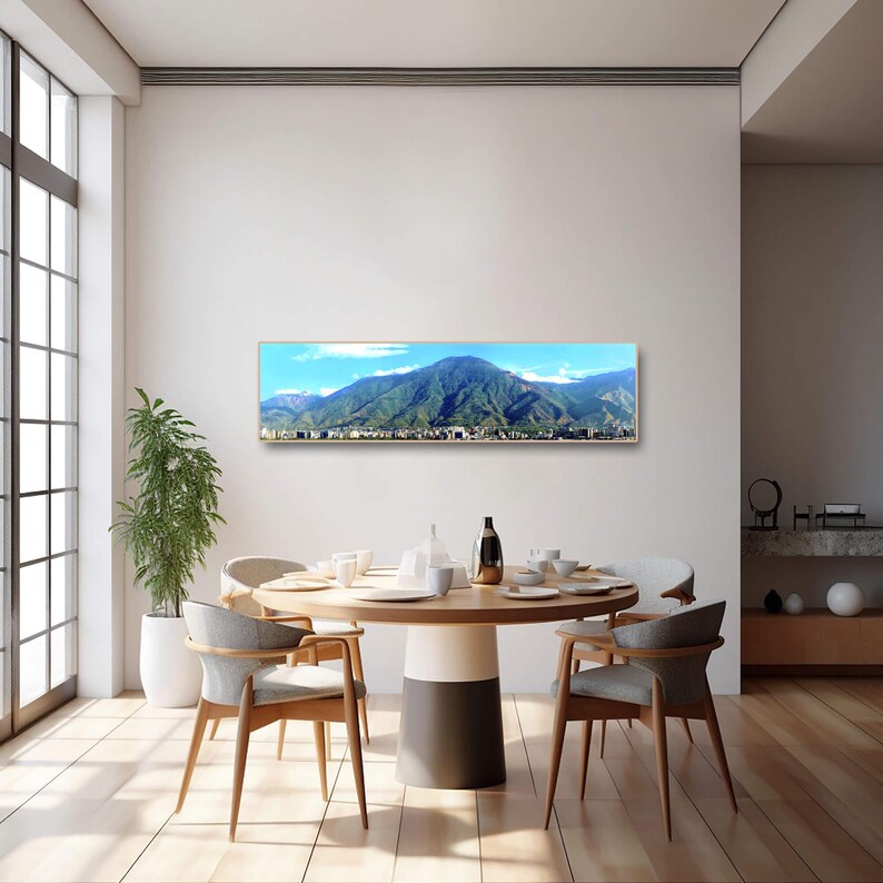 May include: A panoramic photograph of a mountain range with a city in the foreground. The photograph is framed in a gold frame and is hanging on a white wall above a dining table with chairs.