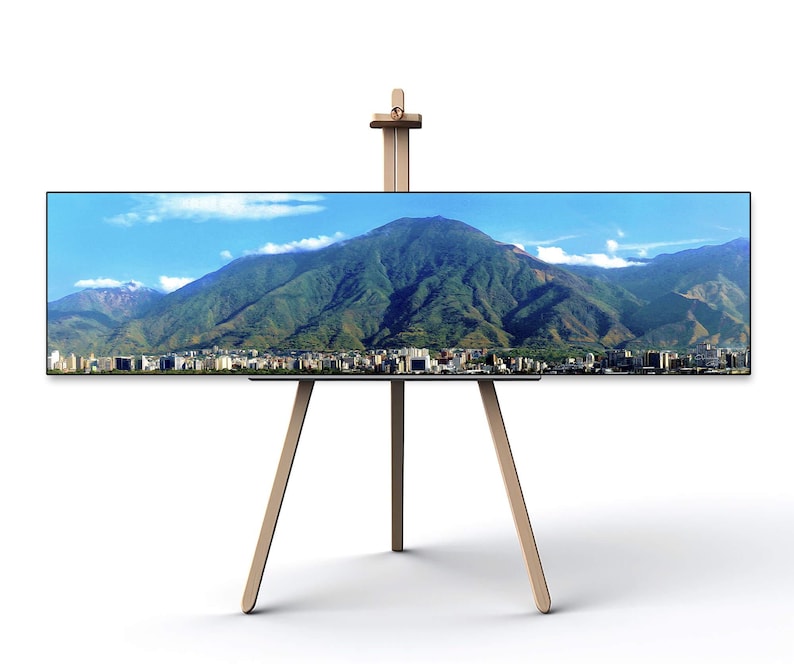 May include: A wooden easel with a wide, rectangular canvas displaying a panoramic photograph of a mountain range with a city in the foreground. The sky is blue with white clouds.