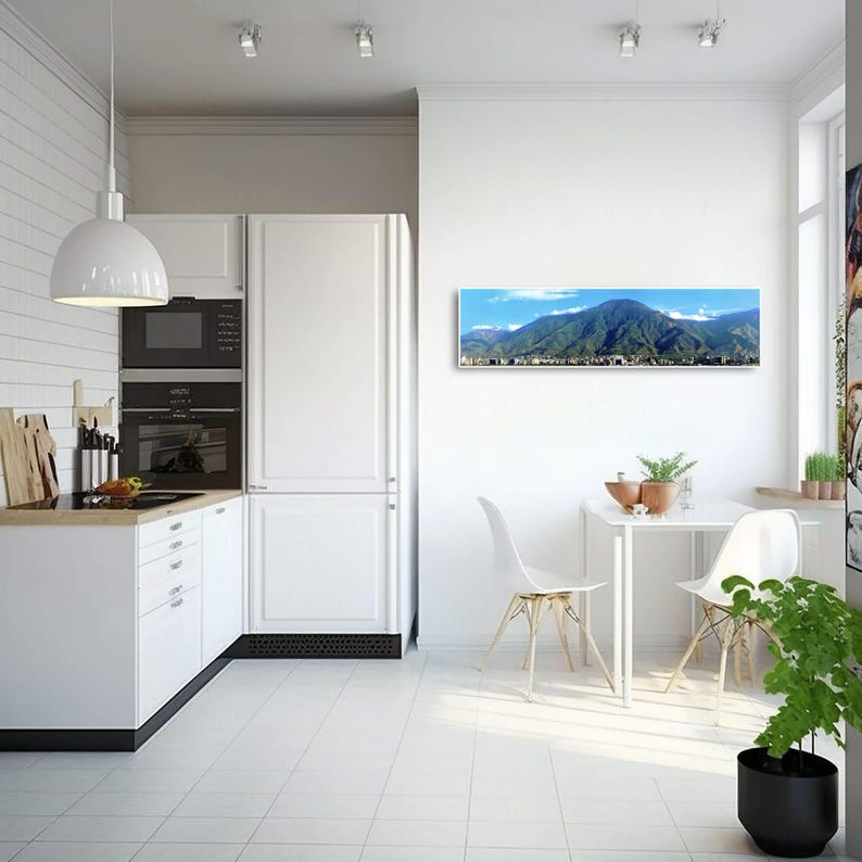 May include: A panoramic photograph of a city nestled in a valley surrounded by mountains. The photo is printed on canvas and framed in a white frame. The photo is hanging on a white wall in a modern kitchen.