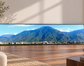 May include: A panoramic wall art print featuring a vibrant landscape of a large, green mountain range towering over a dense city skyline under a bright blue sky with scattered white clouds, all enclosed within a thin, black rectangular frame.