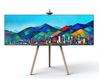 May include: A colorful abstract painting of a city skyline with mountains in the background. The painting is on an easel and is framed in black.