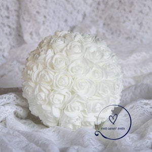 May include: A white round bouquet of artificial roses, arranged in a ball shape, on a white lace background. The bouquet is made of many small white roses, and the background is a white lace fabric. The bouquet is a popular choice for weddings and other special events.