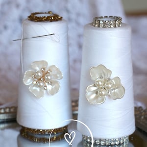 May include: Two white thread spools decorated with pearl and rhinestone embellishments. The spool on the left has a gold beaded trim and a pearl flower. The spool on the right has a rhinestone trim and a pearl flower. The spools are sitting on a mirrored surface with the text "even better event" written on it.