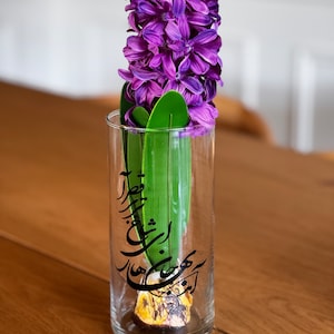 Puede incluir: Un jarrón de vidrio transparente con una flor de jacinto púrpura y hojas verdes. El jarrón tiene escritura caligráfica negra.