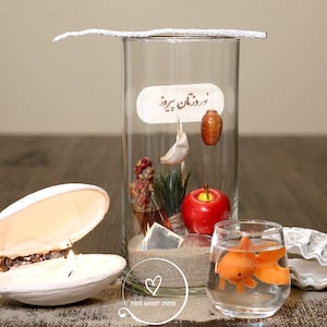 May include: A glass cylinder filled with sand, a red apple, a small plant, a garlic clove, a small brown object, and a label that says "رمضان كريم". There is a white seashell with a candle inside, a glass bowl with a goldfish, and another white seashell on a wooden surface.