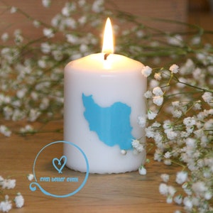 May include: A white candle with a blue outline of the country of Iran on the front. The candle is surrounded by white baby's breath flowers.