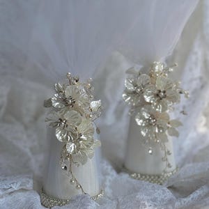May include: Two white cone-shaped wedding decorations with floral embellishments. Each decoration features silver flowers, pearl accents, and a sheer veil. The bases are adorned with a row of small, shiny beads.