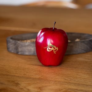 Op de afbeelding: Een rode appel met gouden kalligrafie erop, die op een houten oppervlak ligt.