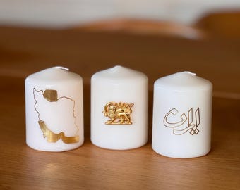 May include: Three white pillar candles with gold designs. The first candle has a gold outline of the map of Iran. The second candle has a gold lion and sun emblem. The third candle has the word "Iran" written in gold Arabic script.