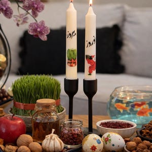 May include: Two lit white candles with Persian script and illustrations stand in black holders. A table is set with a variety of items, including a bowl of nuts, a glass jar, a red apple, and a bowl of goldfish.