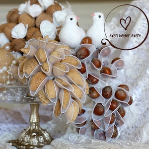 May include: A white cake stand with a gold base holds a wedding centerpiece. The centerpiece features white doves, almonds, and hazelnuts wrapped in white tulle. The text "even better event" is written in brown on a white heart.