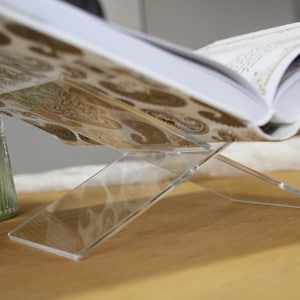 May include: A clear acrylic book stand with a book resting on top. The book has a patterned cover with brown and gold colors.