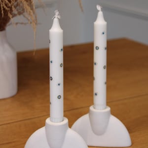 May include: Two white taper candles with blue and gold eye designs, set in matching white holders. The candles stand on a wooden surface, with dried plants in a white vase in the background. Decorative home decor.