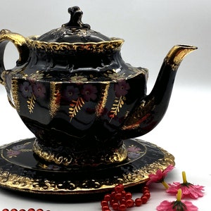 May include: A black teapot with gold accents and floral designs. The teapot has a matching saucer and is decorated with purple and gold flowers.