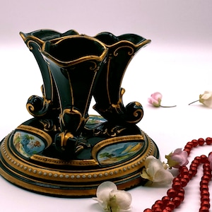 May include: A black and gold ceramic three-horned candlestick with a decorative base featuring painted landscapes. The base is adorned with gold trim and a row of gold dots.
