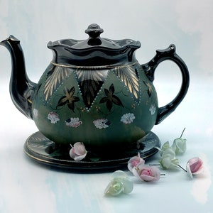 Puede incluir: Una tetera negra y verde con detalles dorados y diseños florales. La tetera está sobre un platillo negro y blanco. Hay varias flores pequeñas de color rosa y blanco esparcidas alrededor de la tetera.