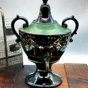 Puede incluir: Una tetera de cerámica negra y verde con detalles dorados y diseños florales. La tetera tiene un pico y una tapa con un pomo. La tetera está sobre una superficie de madera.