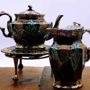 Op de afbeelding: Een zwarte theepot en roomkan set met gouden accenten en bloemmotieven. De theepot heeft een deksel en een handvat, en de roomkan heeft een deksel en een handvat. De set staat op een gouden metalen standaard met drie poten.