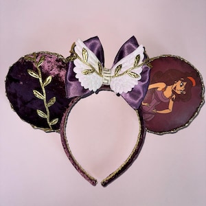 May include: Purple velvet Minnie Mouse ears with a gold trim and a gold leaf design. The bow is purple satin with white wings and gold leaves. The right ear features a printed image of Disney's Megara from Hercules.