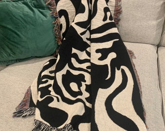 Puede incluir: Manta de tejido negro y blanco con un patrón abstracto ondulado y flecos. La manta está drapeada sobre un sofá.