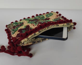 May include: A beige and red fabric clutch with a floral pattern and pom-poms around the edges. A black smartphone is inside the clutch.