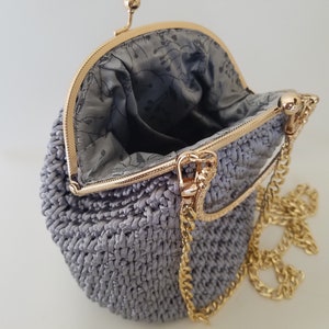 May include: A gray crocheted purse with a gold chain strap and a gold clasp. The purse is open and the inside is lined with a floral pattern.