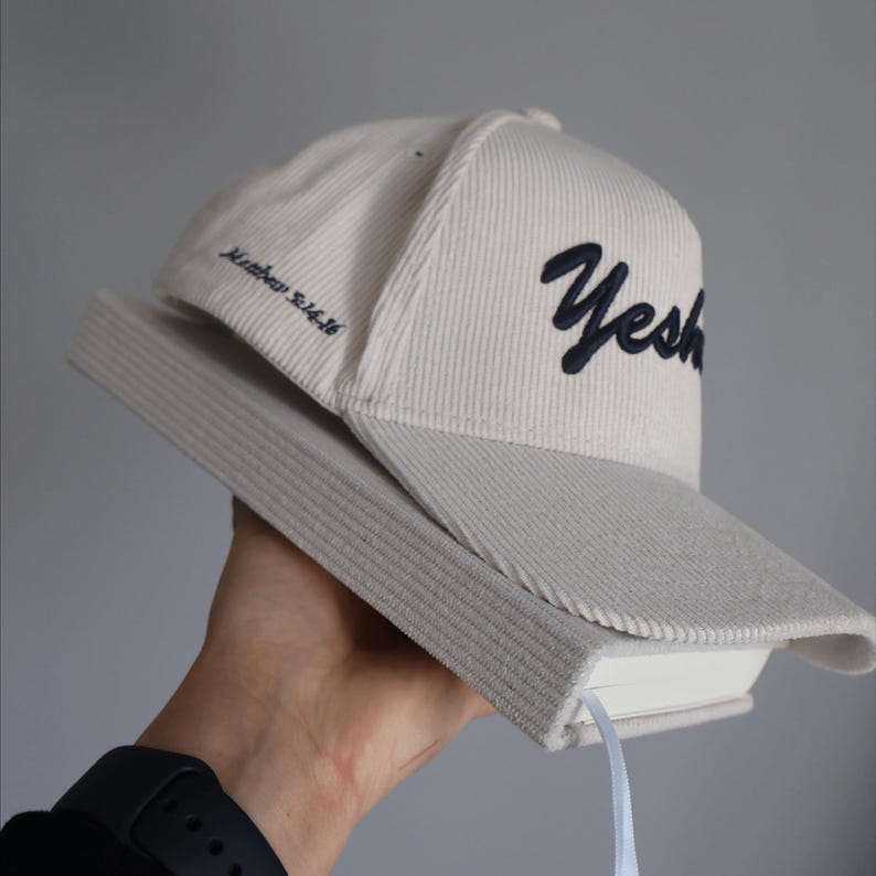 Puede incluir: Una gorra de b&eacute;isbol de pana blanca con un logotipo bordado en negro que dice "S&iacute;" y el texto "Mateo 5:16". La gorra est&aacute; siendo sostenida por una mano y est&aacute; descansando sobre un libro blanco con una cinta azul.