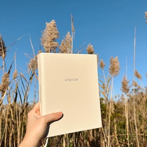 K&ouml;nnte beinhalten: Ein beiger Notizblock mit dem Text "Living in Light" auf dem vorderen Umschlag. Der Notizblock wird in einer Hand vor einem Hintergrund aus hohen, getrockneten Gr&auml;sern und blauem Himmel gehalten.
