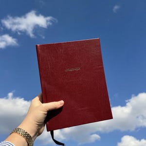May include: A burgundy-colored journal with the words "living for light" embossed in gold. The journal is held against a bright blue sky with fluffy white clouds.