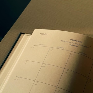 K&ouml;nnte beinhalten: Ein beiger Monatskalender-Planer mit dem Monat M&auml;rz oben. Die Wochentage sind mit Sonntag, Montag und Dienstag beschriftet. Das Zitat "Wenn ihr mich liebt, haltet meine Gebote" - Johannes 14:15 ist oben auf der Seite gedruckt.