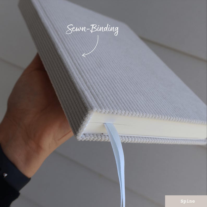 Puede incluir: Un cuaderno gris texturizado con encuadernaci&oacute;n cosida y un marcap&aacute;ginas de cinta azul claro. La encuadernaci&oacute;n del cuaderno es visible, mostrando las p&aacute;ginas blancas en el interior.