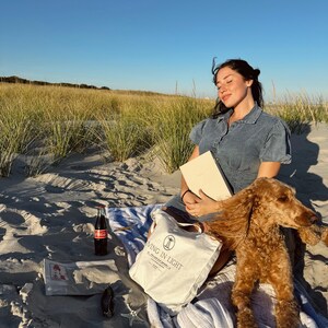 K&ouml;nnte beinhalten: Eine Frau in einem blauen Jeanskleid sitzt am Strand mit einem Hund. Sie h&auml;lt ein Buch und eine wei&szlig;e Einkaufstasche mit dem Aufdruck "Living in Light". Der Hund ist ein brauner Pudel und liegt auf einer wei&szlig;en Decke.