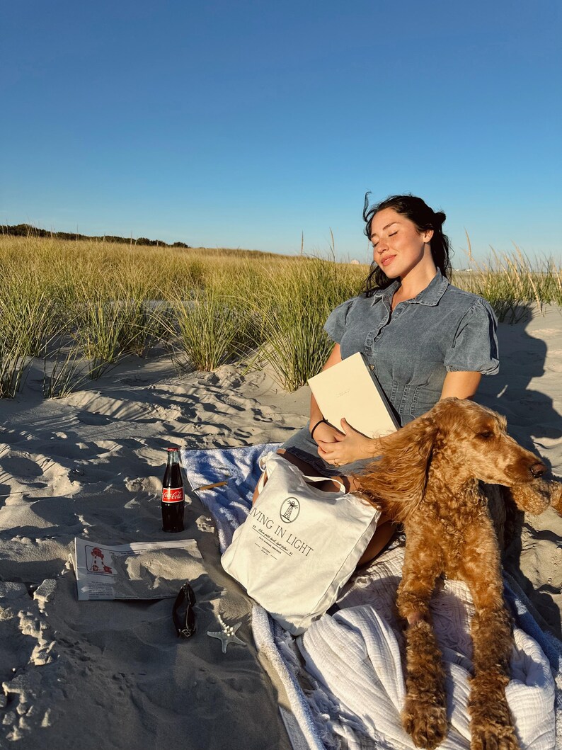 K&ouml;nnte beinhalten: Eine Frau in einem blauen Jeanskleid sitzt am Strand mit einem Hund. Sie h&auml;lt ein Buch und eine wei&szlig;e Einkaufstasche mit dem Aufdruck "Living in Light". Der Hund ist ein brauner Pudel und liegt auf einer wei&szlig;en Decke.