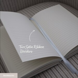 Puede incluir: Cuaderno abierto con p&aacute;ginas rayadas y dos separadores de cinta de sat&eacute;n blanco. El texto "Two Satin Ribbon Dividers" est&aacute; escrito en la p&aacute;gina. El cuaderno est&aacute; sobre una superficie de tela a rayas.