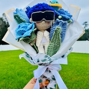 May include: A crocheted bouquet featuring a blue-haired amigurumi doll wearing sunglasses, surrounded by blue and green crocheted flowers. The bouquet is wrapped in white paper with a green leaf pattern and tied with a white ribbon.